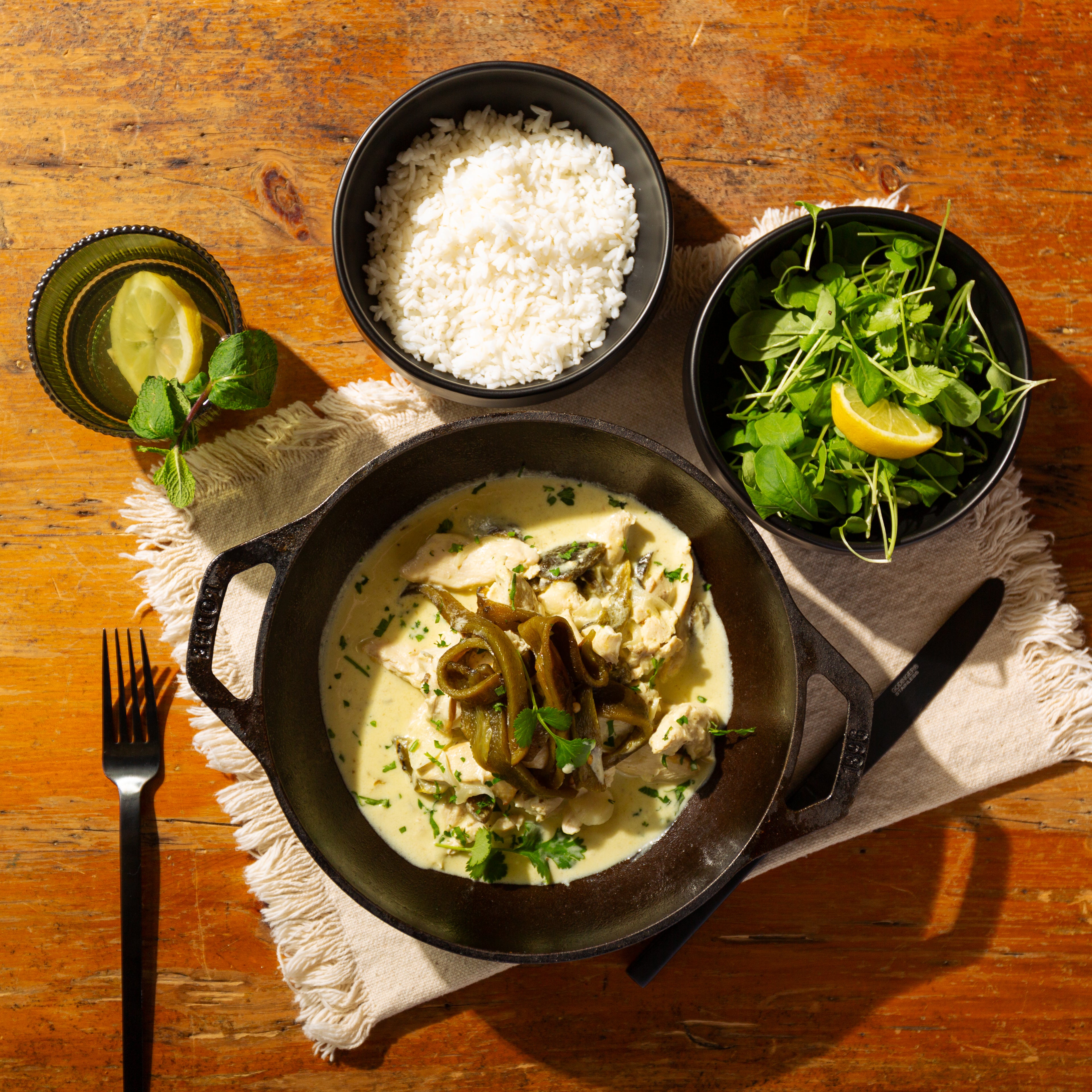 Dish of creamy chicken with green herbs, accompanied by rice and salad on a wooden table.