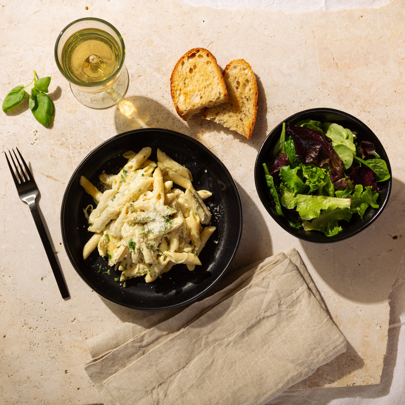 Dinner setting with pasta, salad, bread, and a glass of white wine on a stone surface.