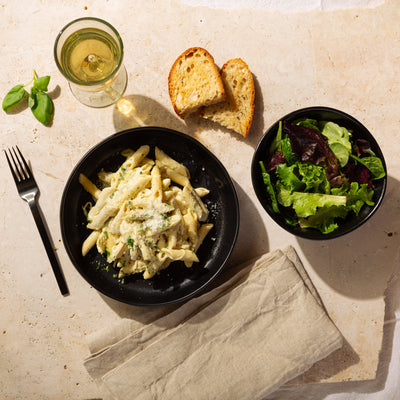 Dinner setting with pasta, salad, bread, and a glass of white wine on a stone surface.