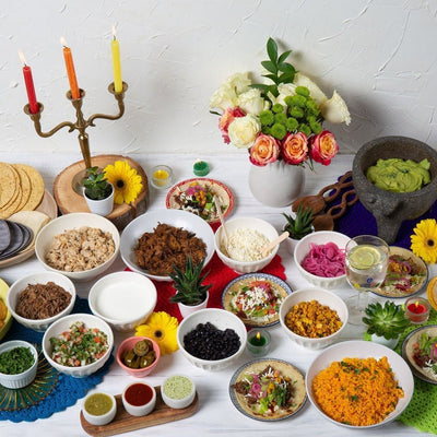 A festive taco bar featuring a variety of prepared dishes including beef Colombian empanadas, corn tortillas, tostadas, chips, fresh cheese, guacamole, pico de gallo, beans, pickled onions, spicy Latin dip, tomatillo sauce, corn, crème fraîche, chipotle sauce, and jalapenos, arranged on a table with decorative elements like candles and flowers in the background.