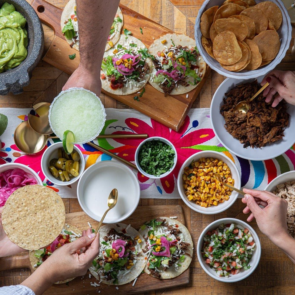 A variety of taco toppings and assembled tacos on a table with people's hands reaching for different ingredients, no specific product to identify.